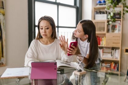 Mamá revisa la tablet mientras su chiquilla con celular conversa sobre límites; escena de niños y smartphones en el hogar